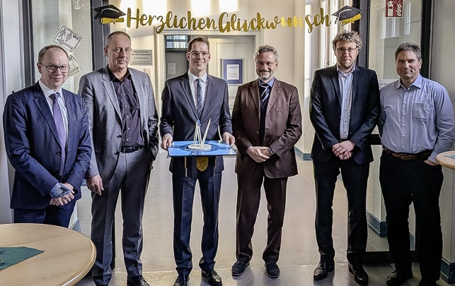 ### Alt-Text**Gruppe Männer in Anzügen bei einer Preisverleihung, Banner 'Herzlichen Glückwunsch' im Hintergrund**