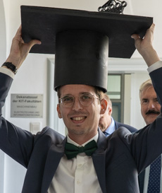 Person hält übergroßen Doktorhut in einer feierlichen Umgebung hoch.