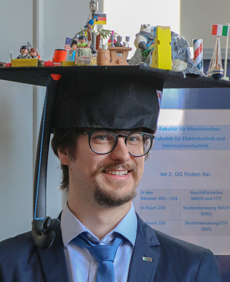 Mann mit Doktorandenhut, dekoriert mit Miniaturmodellen und Flaggen.