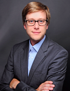 Man in a suit with glasses, arms crossed, against a dark background.