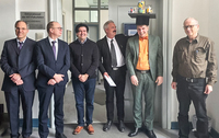 Sechs Männer, einer trägt einen Doktorhut mit Spielzeug, in einem Büroflur.