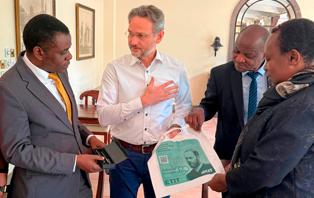 Vier Personen in eleganter Kleidung stehen in einem Raum; eine Person zeigt Geldschein in einer Tüte.