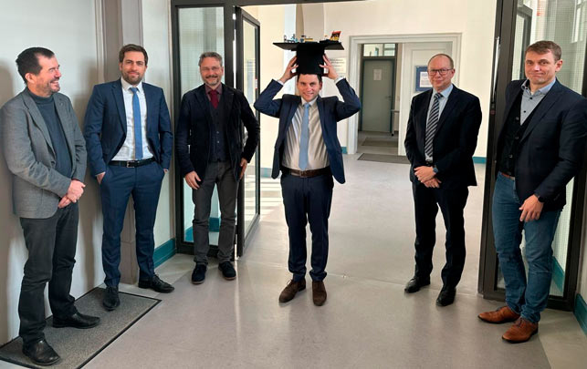 Gruppe von sieben Männern in Anzügen steht in Flur, einer hebt ein Objekt über dem Kopf.